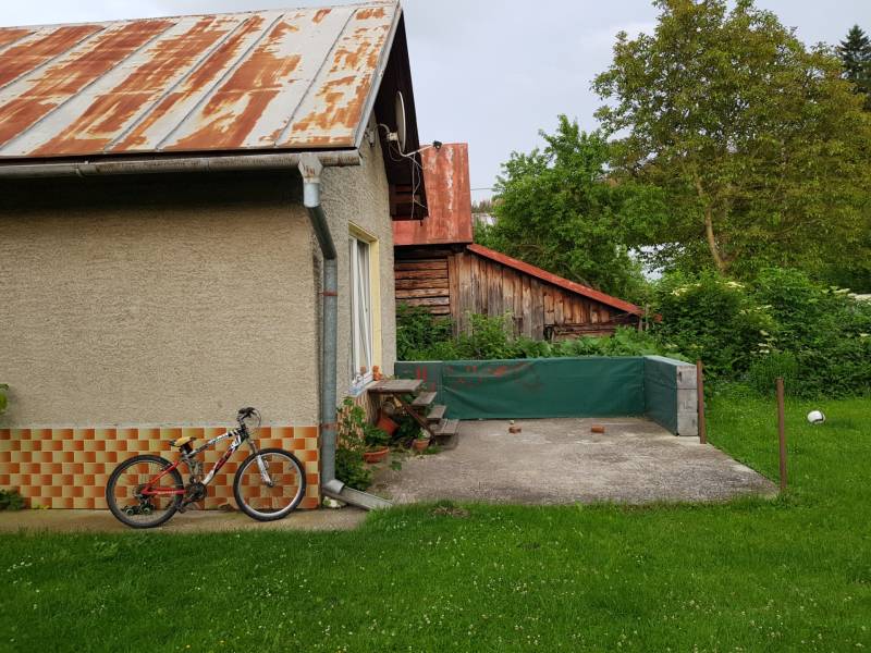 PREDAJ – Rodinný dom s druhou samostatnou stavbou, garážou  - Rodinný dom v Blatnici so záhradou, bicykel opretý o stenu, viditeľný drevený prístrešok.