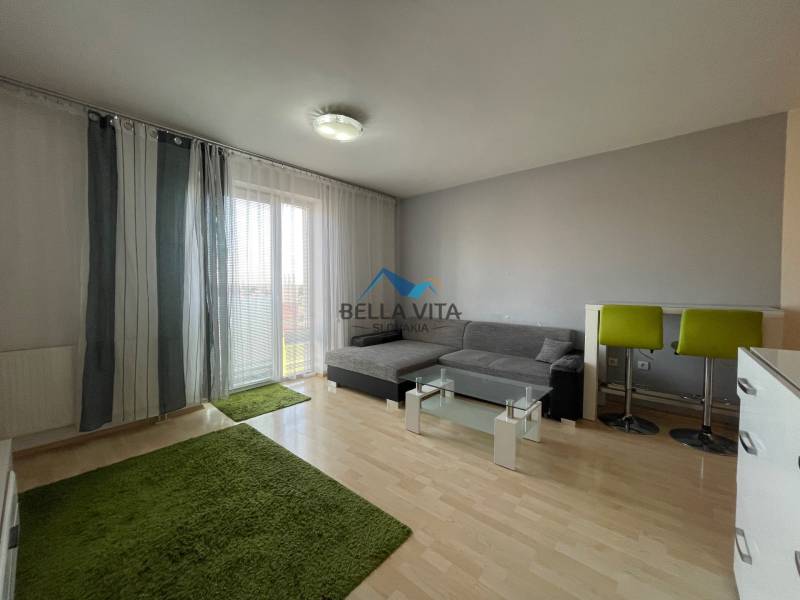 Prenájom 1 izbový byt v Galante, Hodská - Obývacia časť v 1-izbovom byte so sedačkou, zelenými kobercami a podlahou s dreveným dekorom.