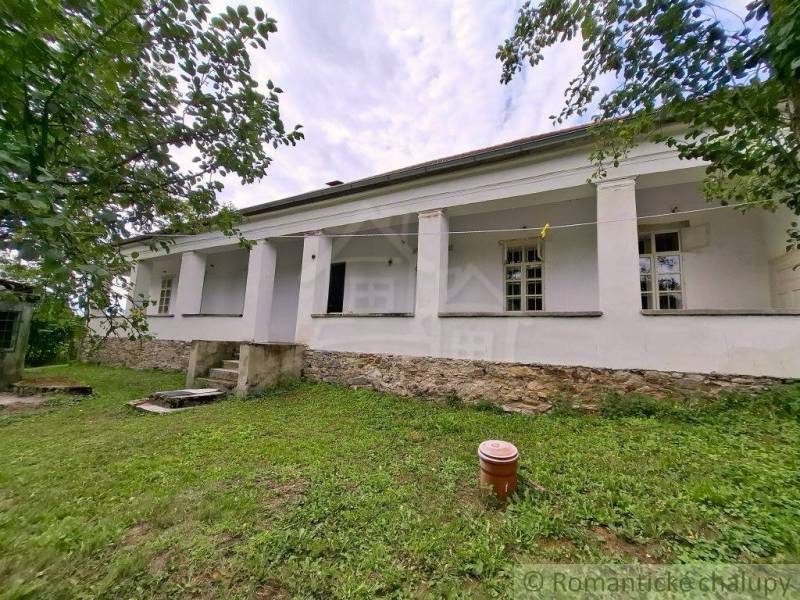 Unikátny meštiansky dom s veľkou záhradou – história, štýl a potenciál - Rodinný dom v Dobroči, biela fasáda, kamenný základ, obklopený zeleňou a stromami.