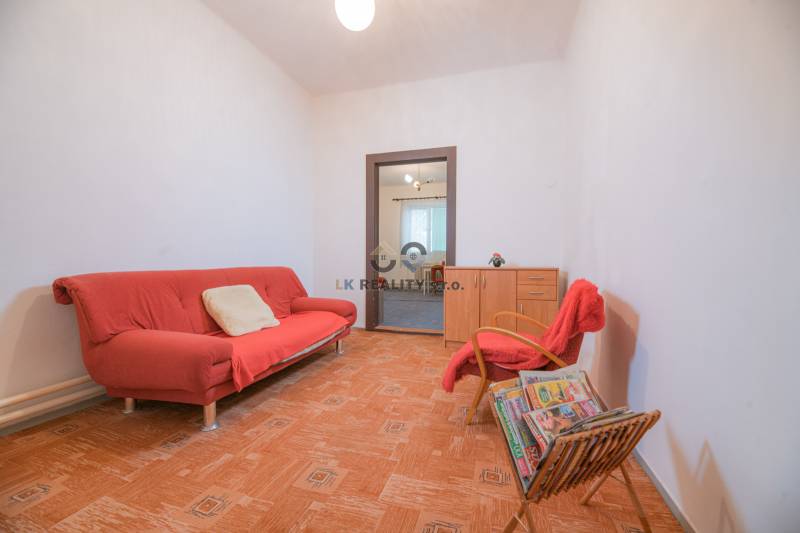 Na predaj rodinný dom Trnava, Zelenečská ulica, pozemok 1085 m2 - Obývacia izba s červeným gaučom, kreslom, časopismi a podlahou s dreveným dekorom v rodinnom dome.