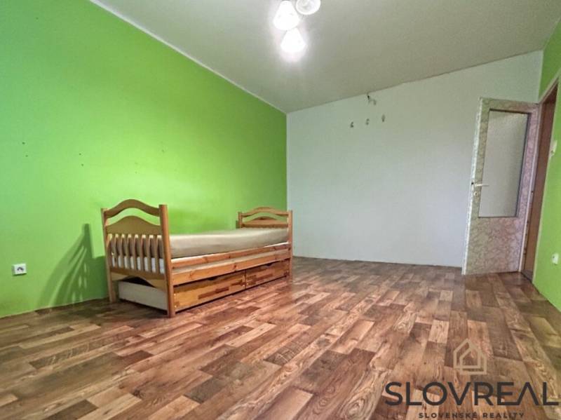 Na predaj 2i byt Levice - Izba s posteľou v 2-izbovom byte, podlaha s dreveným dekorom, zelené steny.