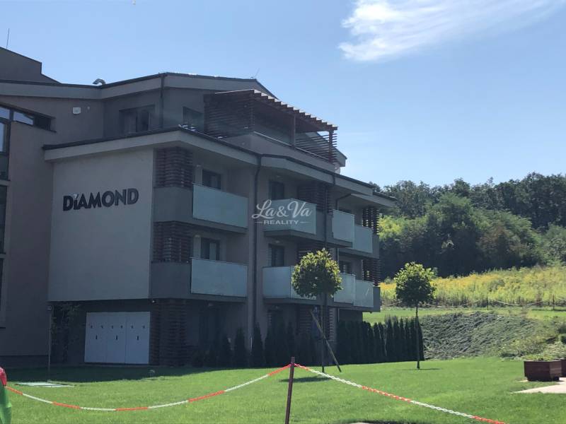 Na predaj apartmán s veľkou terasou Podhájská. - Budova 2-izbového bytu v Podhájskej obklopená zeleňou a bezoblačným nebom.