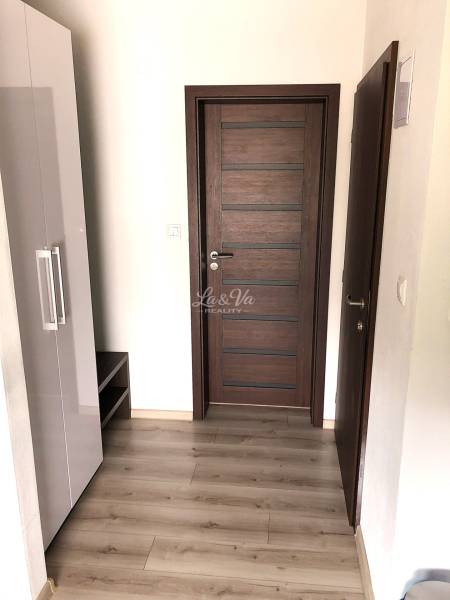 Na predaj apartmán s veľkou terasou Podhájská. - Garsónka s podlahou s dreveným dekorom, tmavými dverami a bielou skriňou.