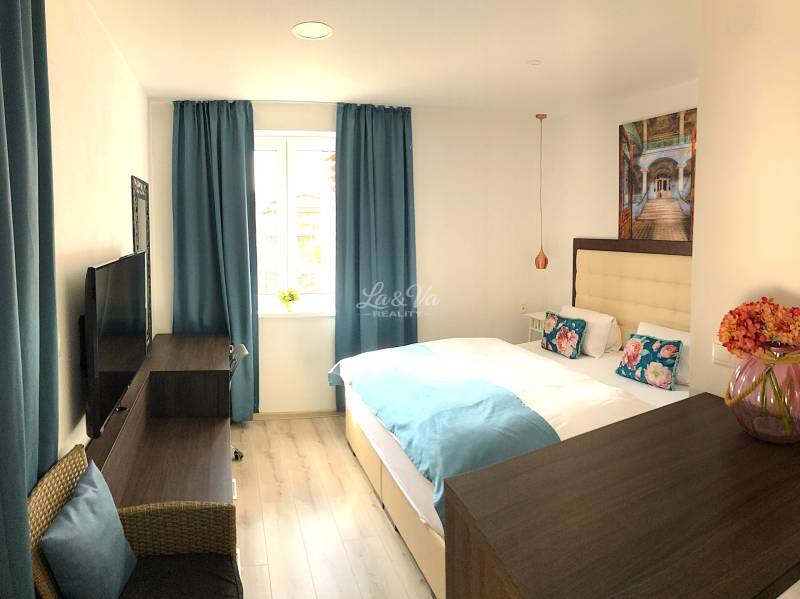 Na predaj štyri apartmány na jednom poschodí Podhájská - Svetlá izba v 2-izbovom byte s modrými závesmi a podlahou s dreveným dekorom.