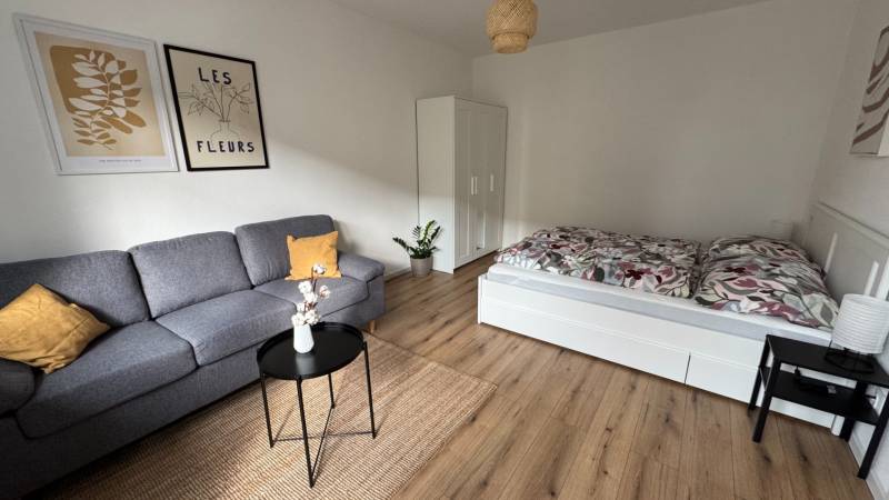 Exkluzívna ponuka moderného 1-izbového bytu s balkónom v meste Vráble - Obývacia izba v 1-izbovom byte s pohovkou, posteľou a podlahou s dreveným dekorom.