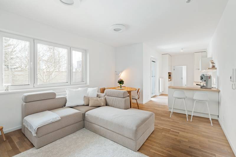 NUERA | PREDAJ 4i byt s predzáhradkou pri lese + 2x parking , BAIV - Svetlá obývacia izba s kuchynským kútom, pohovkou a podlahou s dreveným dekorom.