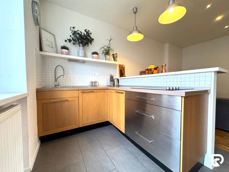 Tichý 2i byt v TOP Lokalite Nív | Trenčianska, BA - Kuchyňa v 2-izbovom byte, sivé dlaždice, drevené skrinky, závesné svetlá, rastliny na poličke.