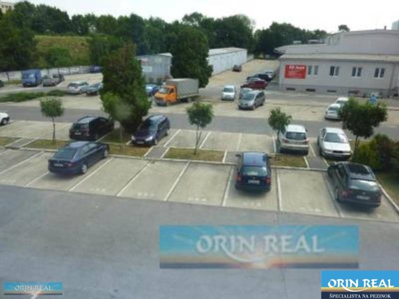 PRENÁJOM KANCELÁRSKÝCH PRIESTOROV NA RAČIANSKEJ ULICI - Parkovisko na ulici Račianska pri budove s kanceláriami, obklopené zeleňou a autami.