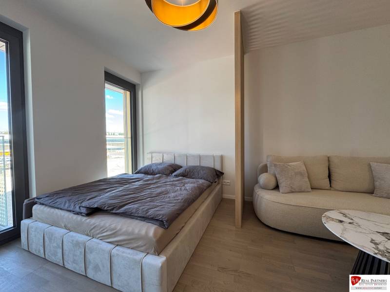 Prenájom 1-izbový byt s balkónom, METROPOLIS, Bratislava- Staré Mesto - Spálňa 1-izbového bytu s posteľou, pohovkou a podlahou s dreveným dekorom.