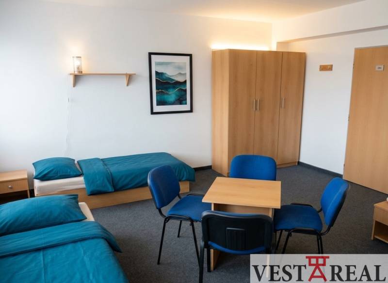 Na prenájom zrekonštruovaná ubytovňa s garážami a skladovacími kobkami - Izba s dvoma posteľami, modrá posteľná bielizeň, skriňa, stôl a štyri stoličky.