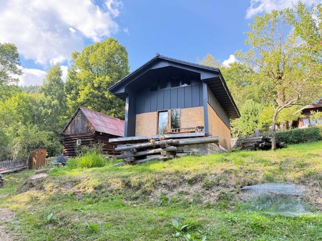 Chata pred dokončením, pozemok 770 m2, Horná Maríková - Chata v Hornej Marikovej, obklopená prírodou a drevenými stavbami, za slnečného dňa.