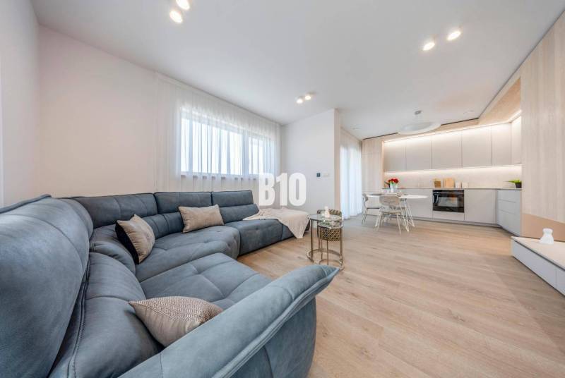 LUXUSNÝ 3-IZBOVÝ BYT NA MIERU V NADČASOVEJ NOVOSTAVBE NA ZAČIATKU KOLI - Obývacia izba s veľkou sedačkou a kuchynským kútom, podlaha s dreveným dekorom.