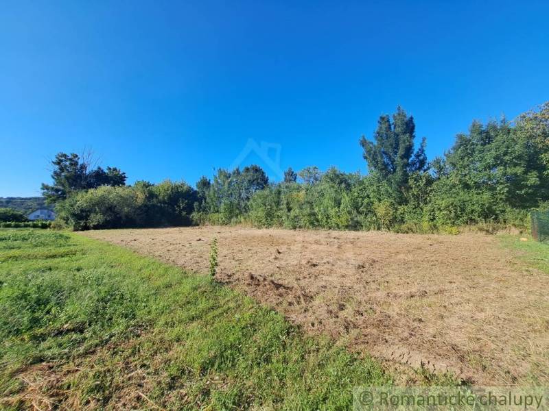 Stavebný pozemok pri Nitre so stavebným povolením v tichej lokalite - Pozemok v Hosťovej na bývanie obklopený zeleňou a modrou oblohou.