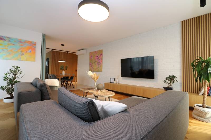 NUERA︱Penthouse s terasou s krásnymi výhľadmi v Dúbravke - Obývacia izba v 4-izbovom byte s podlahou v drevenom dekore, sivou sedačkou a rastlinami.