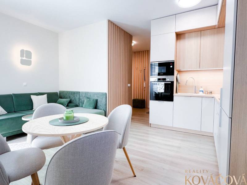 Na predaj 3 izbový byt s terasou, Trenčín, ul. Saratovská - Obývacia izba a kuchyňa v 3-izbovom byte s podlahou s dreveným dekorom a zeleným gaučom.