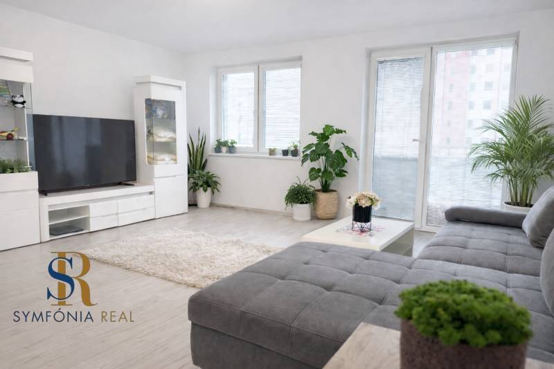 NOVOSTAVBA (2019) + PARKOVANIE | 2-izbový byt 69 m² – Bratislava - Obývacia izba v 2-izbovom byte s rastlinami a nábytkom na podlahe s dreveným dekorom.