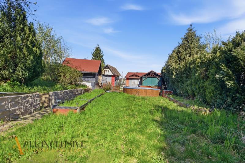 Na predaj záhrada s domčekom, Kráľová nad Váhom - Záhrady v Kráľovej nad Váhom s chatkou, hustou vegetáciou a trávnikom pod modrou oblohou.