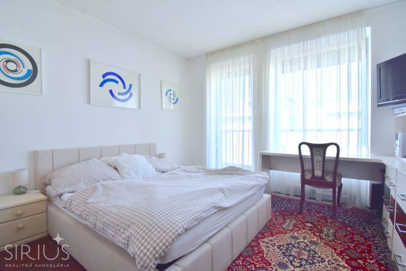 PRI VINOHRADOCH, 3-i byt s veľkými loggiami, 113 m2, RAČANY BIANCO - Spálňa v 3-izbovom byte s posteľou, stolom a umeleckými dielami na stene.