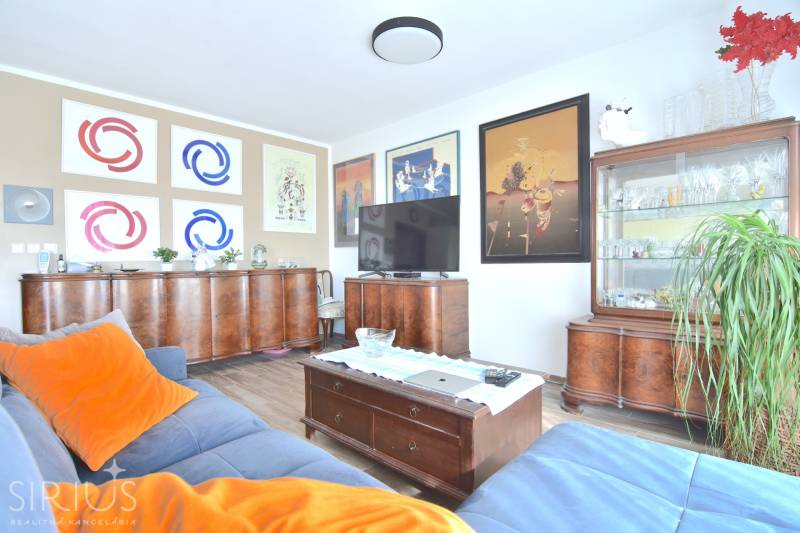 PRI VINOHRADOCH, 3-i byt s veľkými loggiami, 113 m2, RAČANY BIANCO - Obývačka v 3-izbovom byte s dreveným nábytkom, podlahou s dreveným dekorom a obrazmi na stenách.