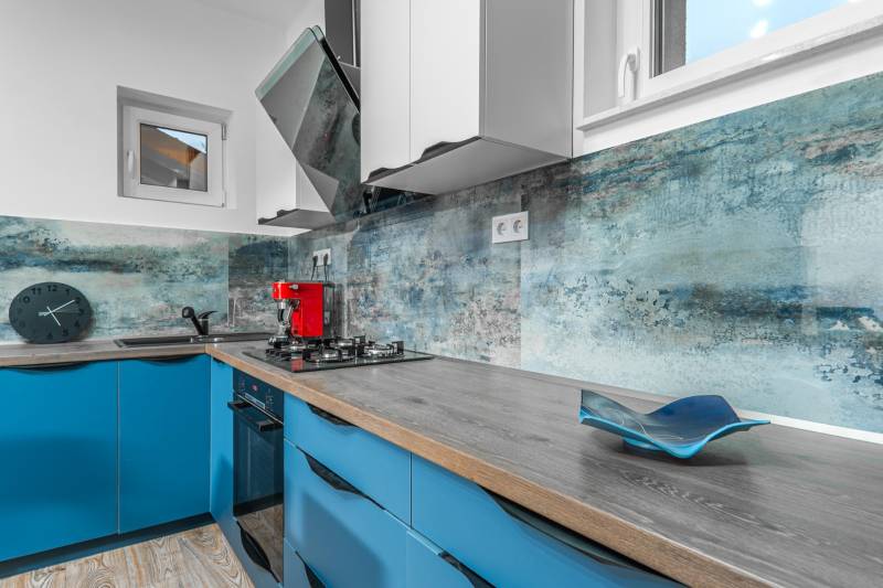 I Predaj štýlového tehlového domu v Rajke pri Bratislave - Kuchyňa s modrými skrinkami, červeným kávovarom a podlahou s dreveným dekorom.