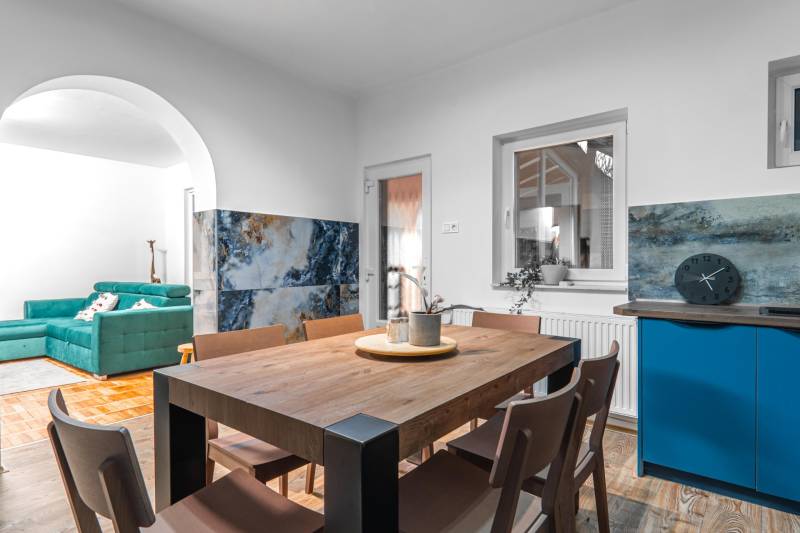 I Predaj štýlového tehlového domu v Rajke pri Bratislave - Jedáleň v rodinnom dome s dreveným stolom, modrou kuchynskou stenou a pohovkou v pozadí.