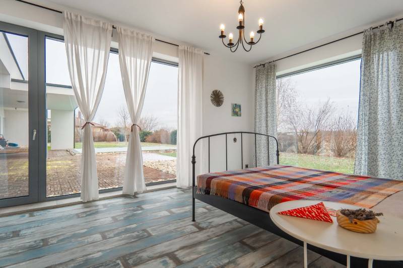 I PREDAJ moderná vila s bazénom a veľkým pozemkom - Spálňa vo vile s vysokými oknami, kovovou posteľou a podlahou s dreveným dekorom.