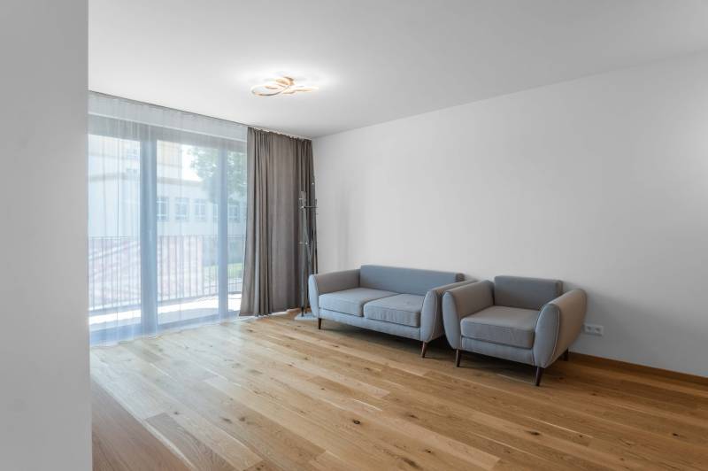 : Nadštandardný 2-i byt v projekte ZWIRN za cenu „štandardu“ - Svetlá obývacia izba so sivou pohovkou, drevenou podlahou a balkónovými dverami v 2-izbovom byte.