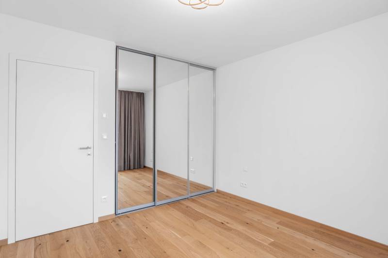 : Nadštandardný 2-i byt v projekte ZWIRN za cenu „štandardu“ - Izba v 2-izbovom byte s bielymi stenami, zrkadlovou skriňou a drevenou podlahou.