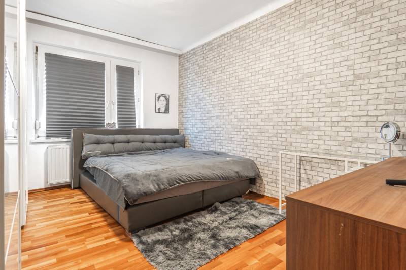 I Na predaj 2-i byt s terasou v Auersthal (Rakúsko) - Spálňa so sivou posteľou, tehlovou tapetou a podlahou s dreveným dekorom v 2-izbovom byte.