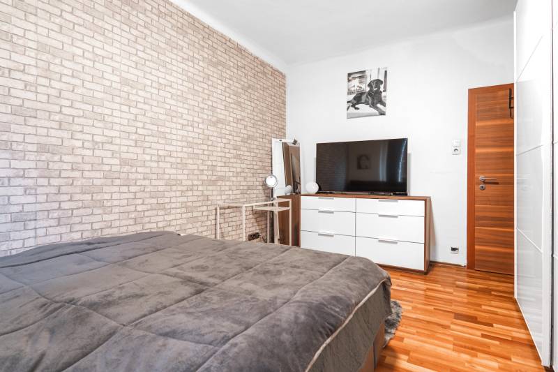 I Na predaj 2-i byt s terasou v Auersthal (Rakúsko) - Spálňa s tehlovou tapetou, televízorom, komodou a podlahou s dreveným dekorom v 2-izbovom byte.