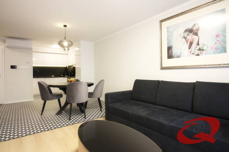 **SKVELÁ INVESTÍCIA**REZIDENCIA OPERA**KÚPELE TURČIANSKE TEPLICE** - Obývačka v 2-izbovom byte s kuchyňou, čierna sedačka, umelecký obraz, podlaha s dreveným dekorom.