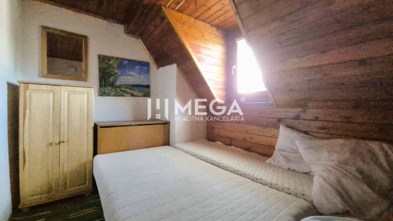 PREDAJ REKREAČNEJ CHATY NA DOMAŠI časť DOBRÁ - Izba v chate s posteľou, drevenými stenami a skrinkou pod šikmým stropom.