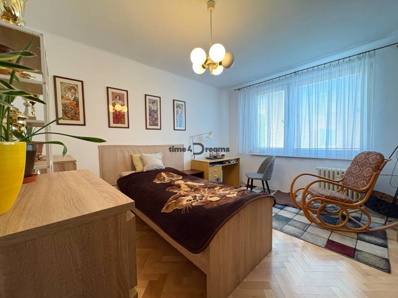 Na predaj: 4 izb.byt + BALKON + ŠATNIK + ŠPAJZA - Spálňa v 3-izbovom byte s podlahou s dreveným dekorom, nábytkom a hojdacím kreslom.