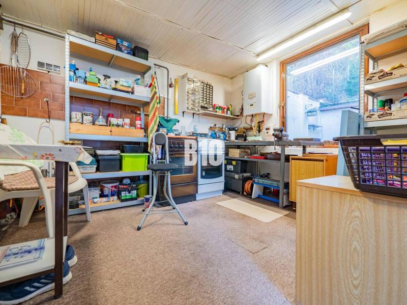 POD HRADOM bývanie alebo podnikanie - Dielňa v rodinnom dome s regálmi, náradím a stolom. V okne je vidieť exteriér.