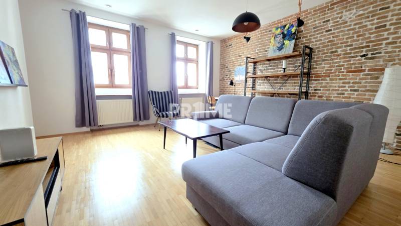 JEDINEČNÝ 2i byt, REKONŠT., 2 X .SAMOST.IZBA, Hviezdoslavovo nám.  - Obývacia izba s tehlovou stenou, sivou sedačkou a podlahou s dreveným dekorom v 2-izbovom byte.