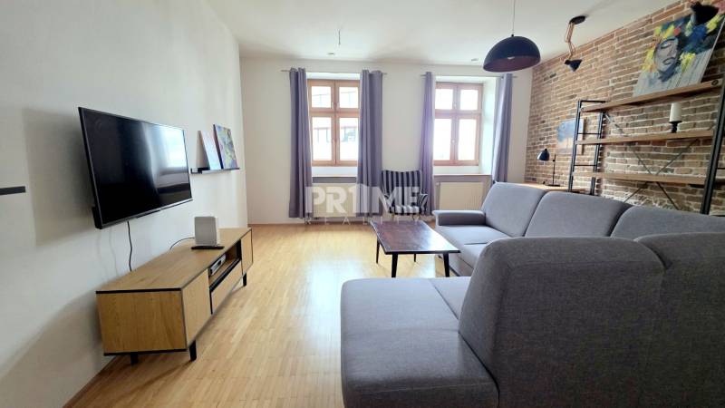 JEDINEČNÝ 2i byt, REKONŠT., 2 X .SAMOST.IZBA, Hviezdoslavovo nám.  - Obývacia izba v 2-izbovom byte, sivá sedačka, televízor, tehlová stena, podlaha s dreveným dekorom.