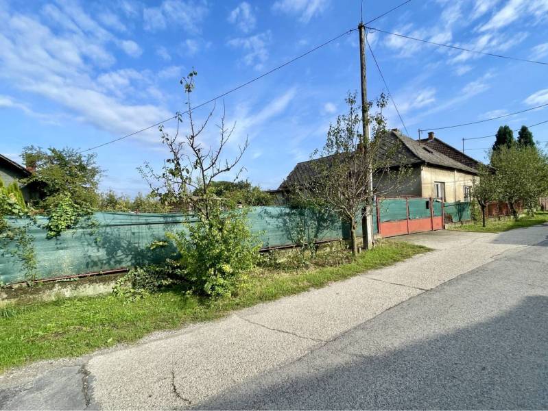 JAHODNÁ – SLNEČNÝ POZEMOK S STARÝM RODINNÝM DOMOM NA PREDAJ - Rodinný dom v Jahodnej s oplotením a stromami popri neuvedenej ulici za slnečného dňa.