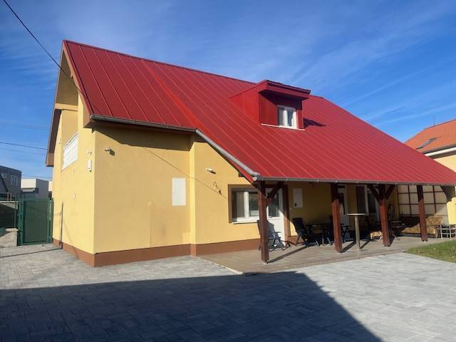 Apartmány na prenájom. - Rodinný dom s červenou strechou a terasou, popisuje vidiecke prostredie a budovu.