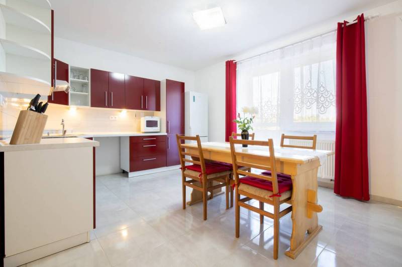 Dvojpodlažný rodinný dom s veľkým pozemkom na predaj v obci Jahodná - Kuchyňa so svetlým stolom, červenými skrinkami a závesmi, ideálna pre rodinný dom.