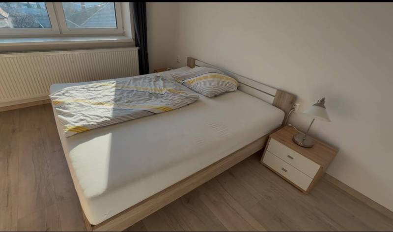 Prenájom 2-izbového bytu, Dunajská Streda - Manželská posteľ vedľa nočného stolíka s lampou, podlaha s dreveným dekorom.