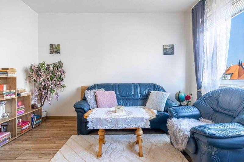 ☎️ 0918197701- PREDAJ - Nebytových priestorov Dunajská Streda - Modrý kožený gauč s dekoratívnymi vankúšmi, knihovna a podlaha s dreveným dekorom.