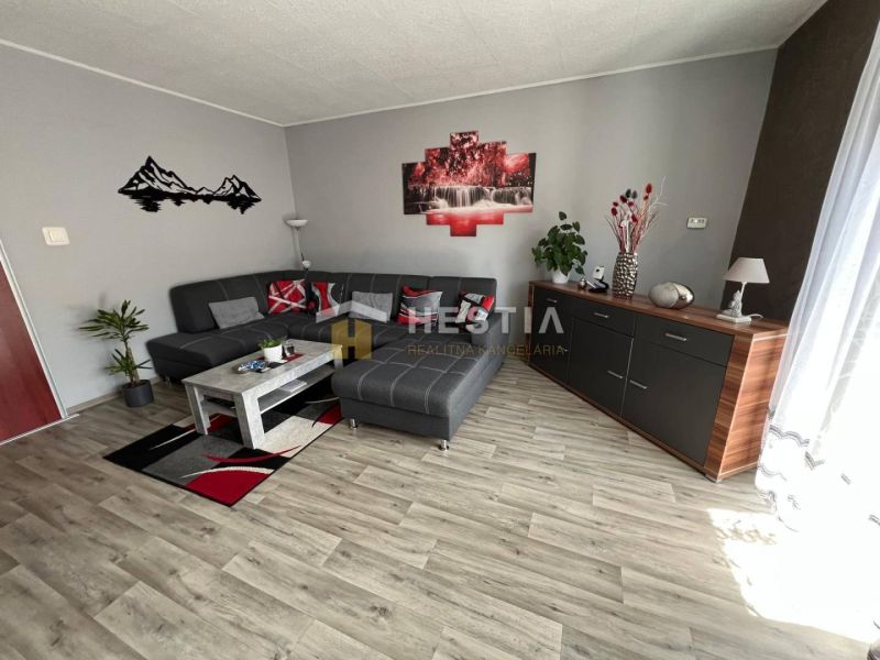 NOVINKA Priestranný 3-izbový byt (99 m²) s pozemkom, terasou, 2 balkón - Obývacia izba v 3-izbovom byte s podlahou s dreveným dekorom a šedým gaučom.