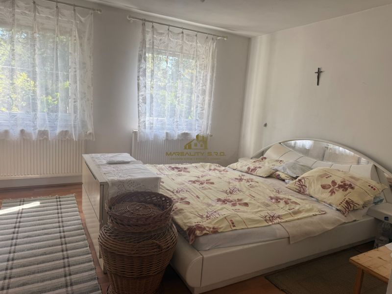 NA PREDAJ celoročná chata – Sekule (Šutrovňa), tiché prostredie - Spálňa v chate s posteľou, kobercom, bielou komodou a závesmi, podlaha s dreveným dekorom.