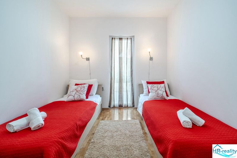 Brač(HR) - Apartmány v 1. rade u mora !!! - Rekreačný apartmán s dvomi posteľami, červenou prikrývkou a podlahou s dreveným dekorom.