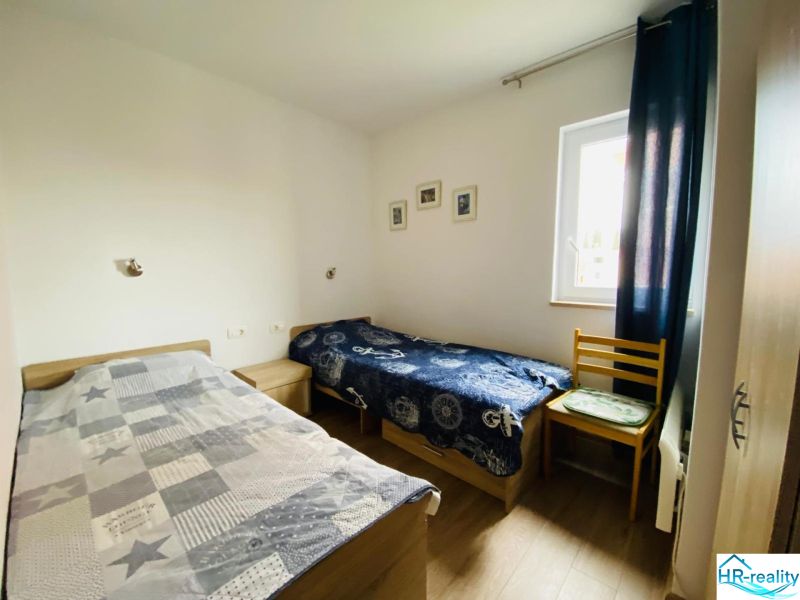 Brač(HR)–Apartmán s výhľadom na more - Dve postele, stolička, okno, obrazy na stenách, podlaha s dreveným dekorom.