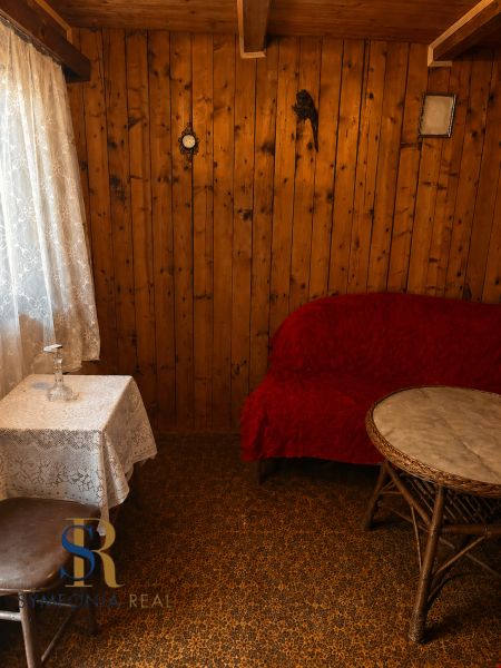 REZERVOVANÉ - Záhrada, ticho, vlastný priestor | pozemok s chatkou - Drevené steny, čipkovaný obrus, červený poťah a okrúhly stolík v Chate.