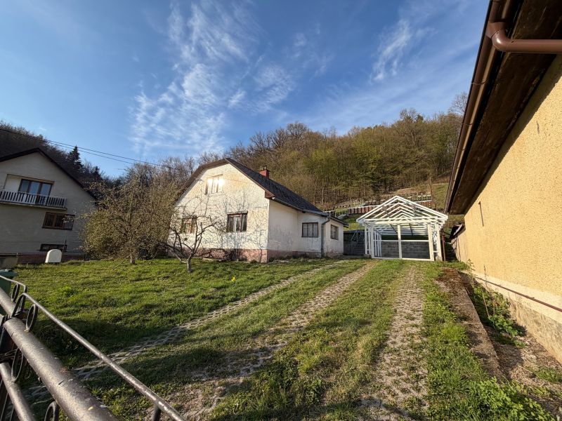 RODINNÝ DOM V PRÍRODE - NA PREDAJ -OBEC ŠTÓS  - Rodinný dom v Štóse obklopený zeleňou a drevenou konštrukciou prístrešku.