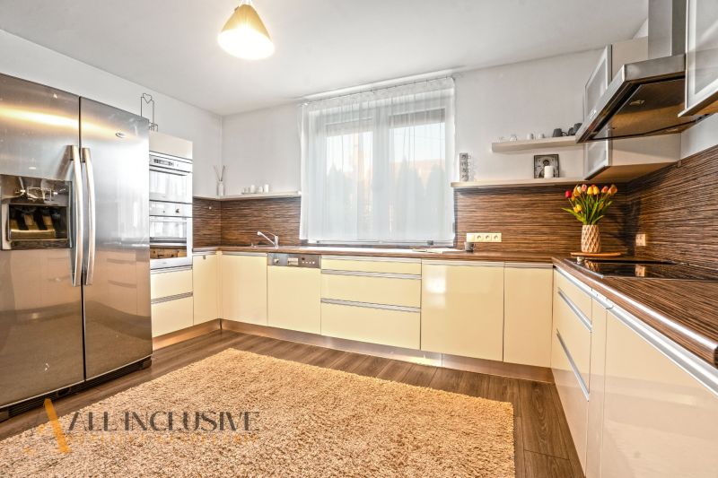 NA PREDAJ 4-IZBOVÝ RODINNÝ DOM V TOMÁŠIKOVE - Kuchyňa v rodinnom dome s béžovým nábytkom, veľkou chladničkou a podlahou s dreveným dekorom.