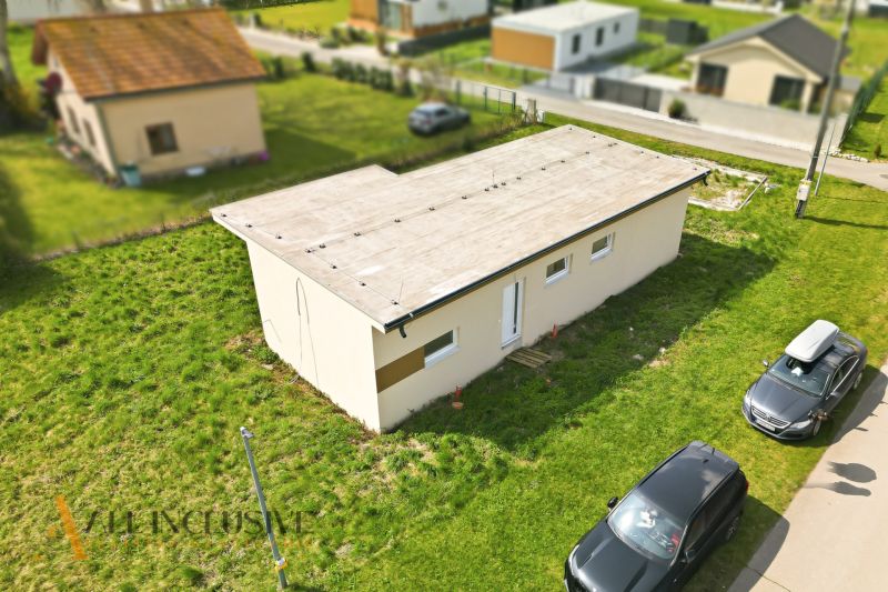 Na predaj rekreačný dom pri jazere, Vojka nad Dunajom - Chata vo Vojke nad Dunajom s rovnou strechou a zaparkovanými autami na trávnatej ploche.