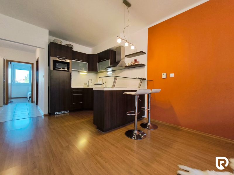 | priestranný | 2i byt s balkónom | parkovaním | Koprivnická - Kuchyňa v 2-izbovom byte s výraznou oranžovou stenou a podlahou s dreveným dekorom.